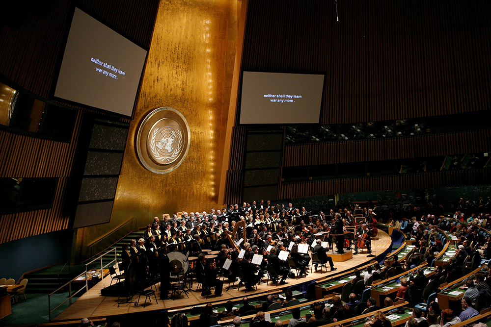 Chor und Orchester auf dem Podium der UN-Generalversammlung in New York, auf den Leinwänden der Text „neither shall they learn war any more“