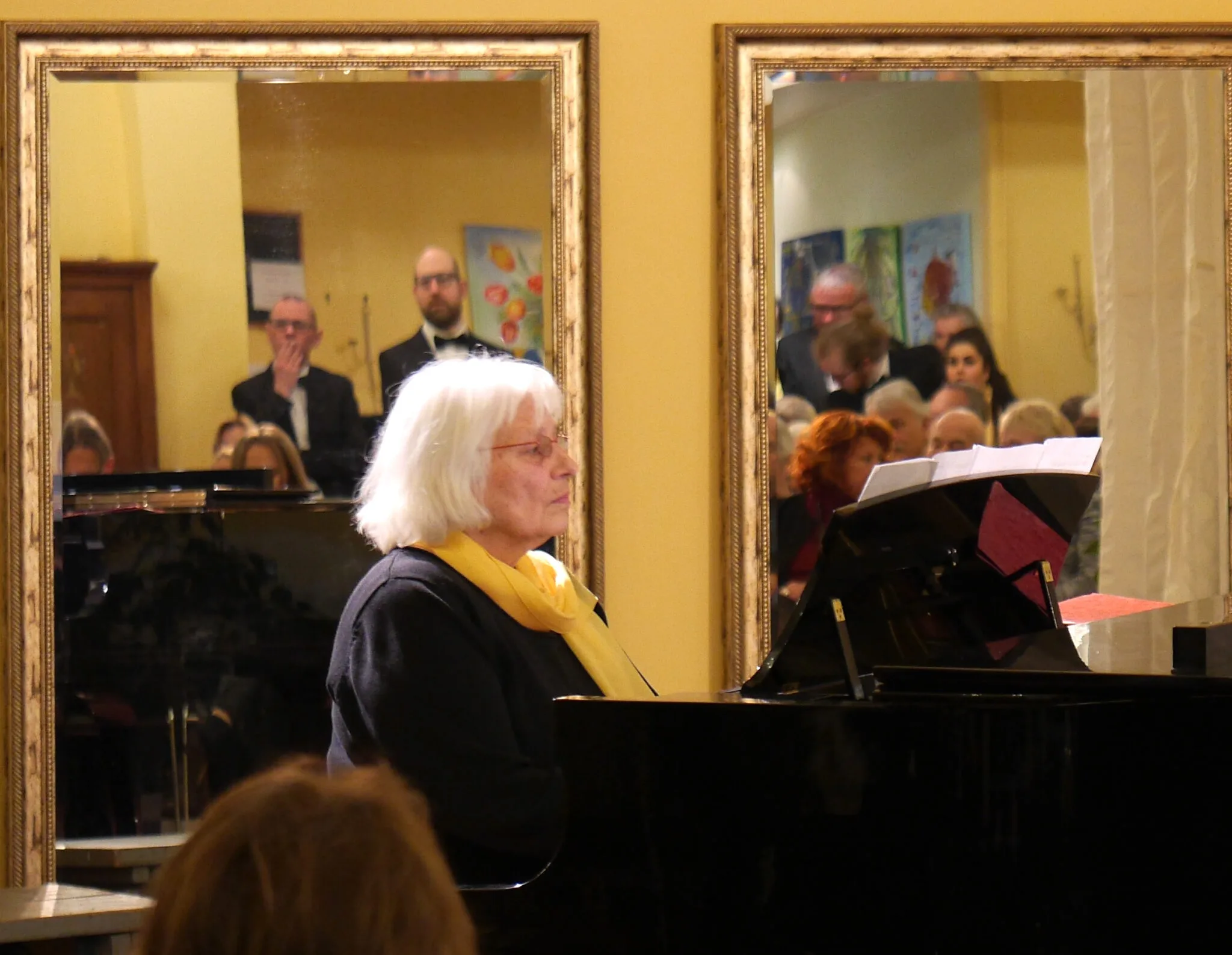 Eine Pianistin mit gelbem Schal am Flügel, im Spiegel dahinter Sänger des Chores und Publikum